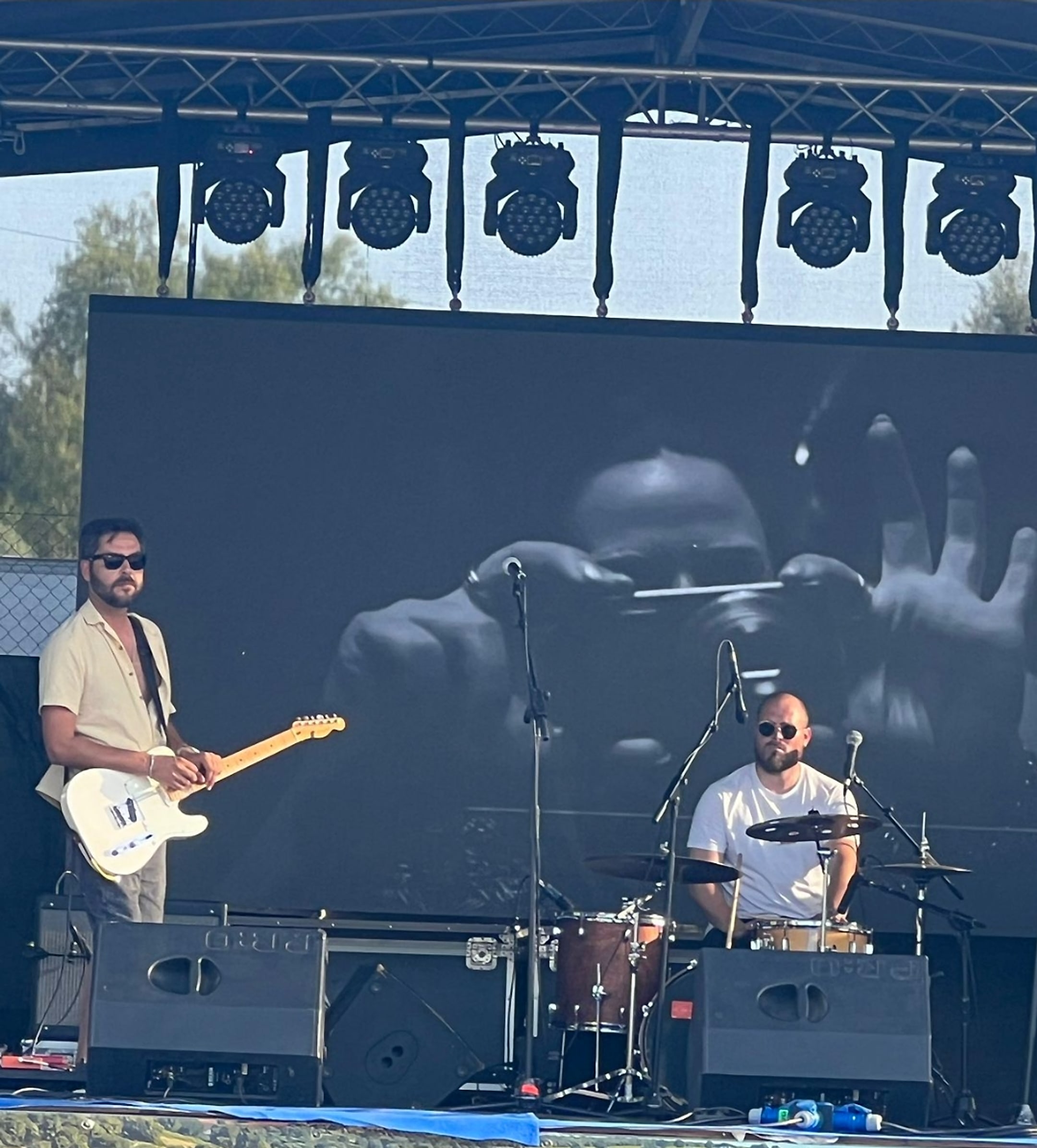 António Roque playing a white electric guitar on an outdoor stage, with a drummer beside him